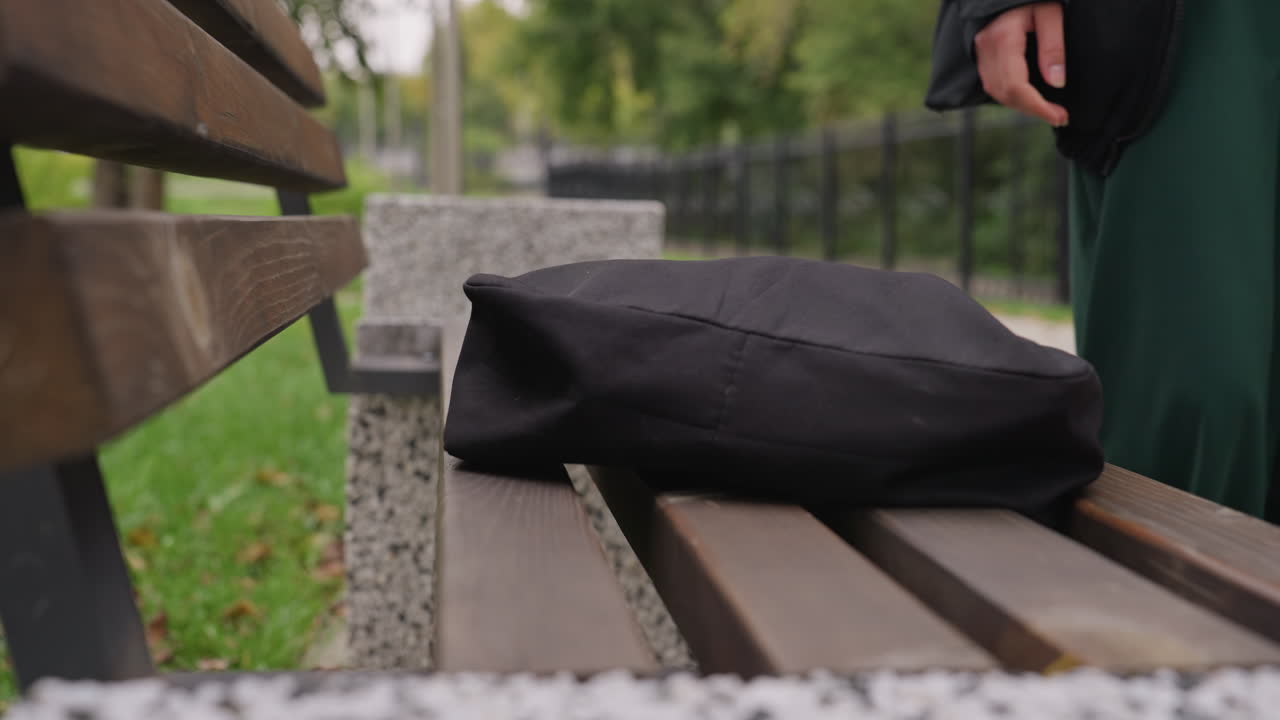 Zaino su una panchina, donna bianca reclinata, sguardo pensieroso verso il cielo, cappotto verde, listelli di legno, sentiero del parco e recinzione sullo sfondo, foglie autunnali sull'erba, messa a fuoco superficiale sulla borsa, atmosfera di solitudine urbana, possibile
