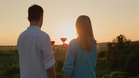 Pareja-Joven-Con-Copas-De-Vino-Tinto-Contemplando-La-Puesta-De-Sol-Sobre-Un-Valle-Pintoresco