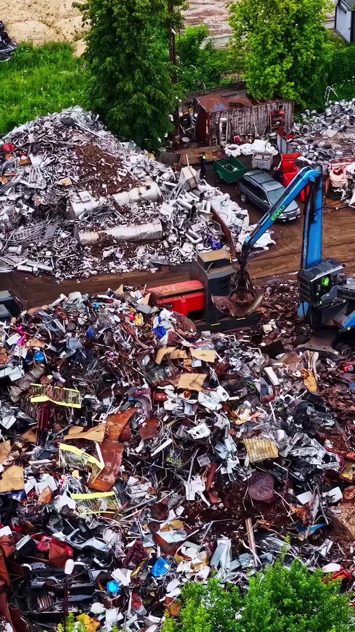 Inquadratura aerea di macchinari per il riciclo che lavorano tra grandi cumuli di rifiuti metallici misti, ripresa verticale con drone
