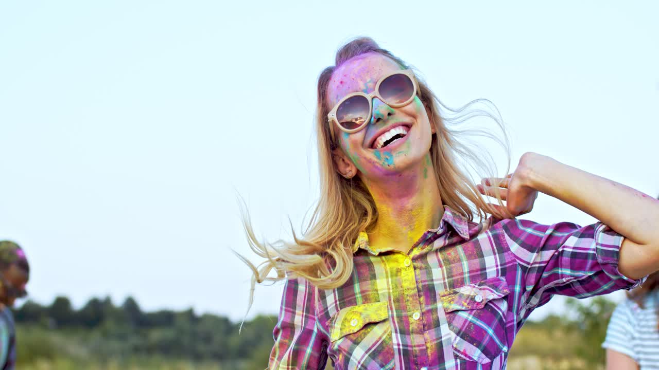 joven mujer rubia atractiva y feliz con gafas de sol saltando, bailando y divirtiéndose con amigos de razas mixtas al aire libre en la celebración de las vacaciones de holi