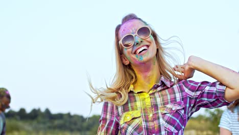 joven mujer rubia atractiva y feliz con gafas de sol saltando, bailando y divirtiéndose con amigos de razas mixtas al aire libre en la celebración de las vacaciones de holi