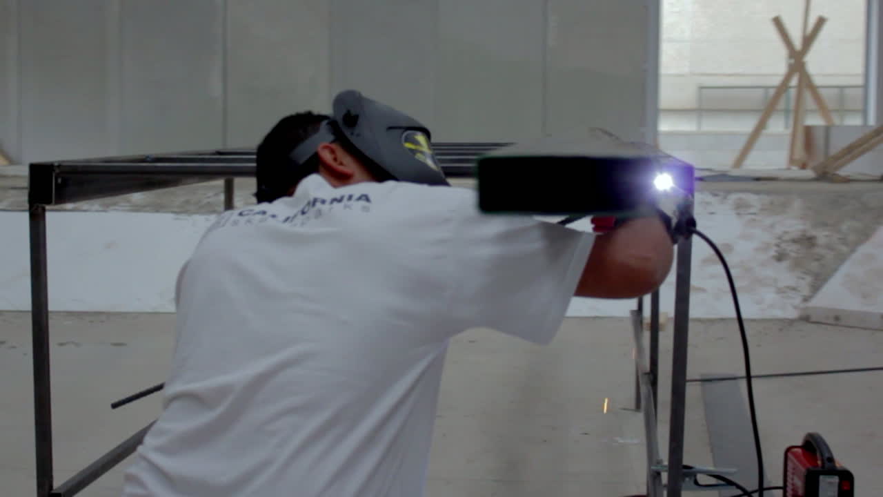 A welder leans in with precision, sparks glowing as metal fuses under intense concentration in a quiet, industrial workspace.