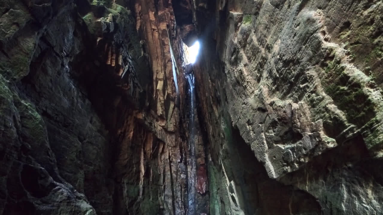 A dramatic view from inside a narrow cave, showing a thin waterfall cascading down dark, mossy stone walls with bright daylight streaming through the opening above