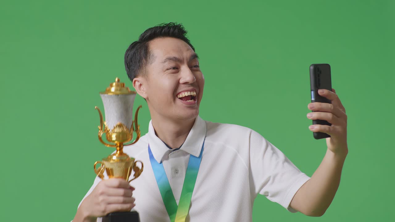 골드 메달을 가진 아시아 남자의 클로즈업은 스튜디오의 녹색 화면 배경에서 스마트폰에서 비디오 통화에 골드 트로피를 보여줍니다.