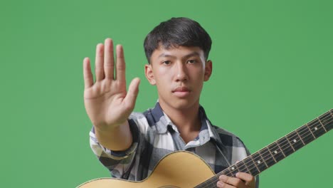 close up of asian teenager with guitar making stop gesture while standing on green screen background