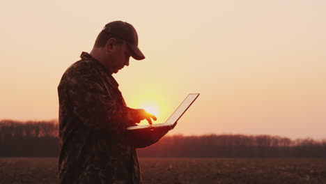 Ein-Bauer-Mittleren-Alters-Benutzt-Einen-Laptop-Bei-Sonnenuntergang-Auf-Einem-Feld-Und-Plant-Die-Aussaat