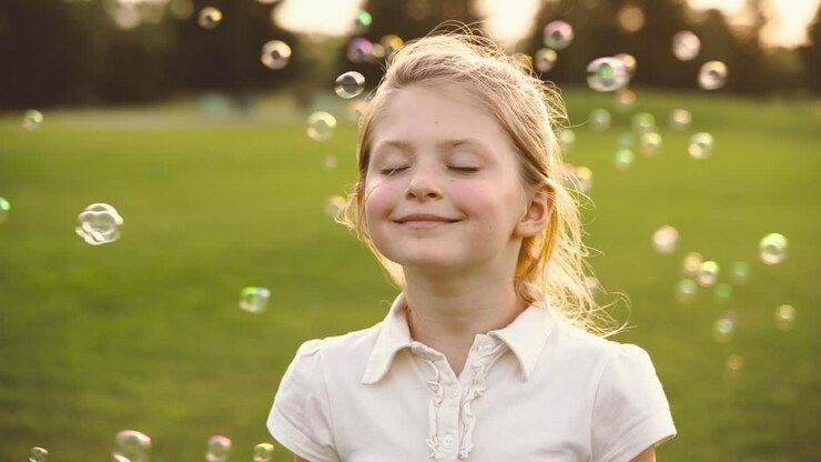 Ritratto di una graziosa bambina bionda sorridente, con gli occhi chiusi, circondata da bolle di sapone nel parco 1