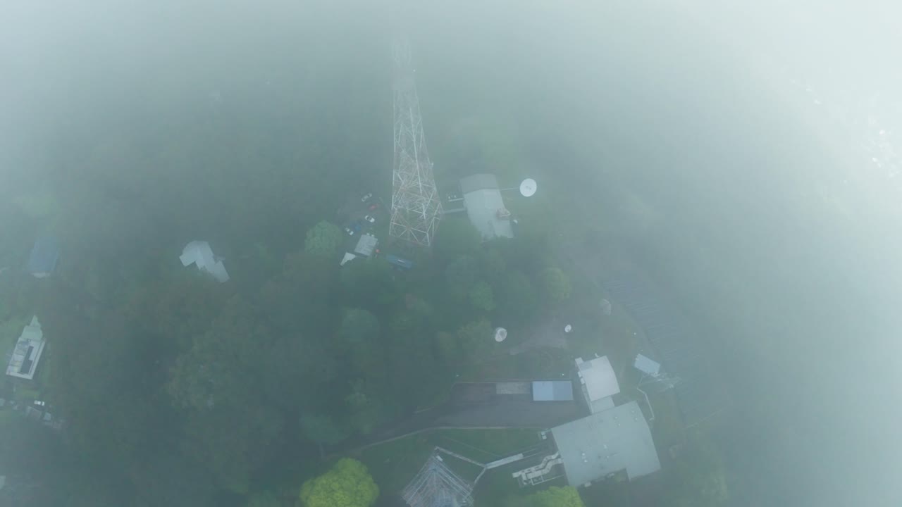 Drone footage capturing a towering communication antenna amidst lush, fog-covered forests. The green landscape and mist create a mysterious, atmospheric scene in a rural setting