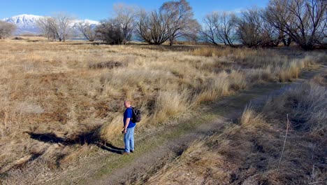 Mature-man-with-a-backpack-out-for-a-hike-and-enjoying-the-snow-capped-mountain-view---aerial-orbit-view