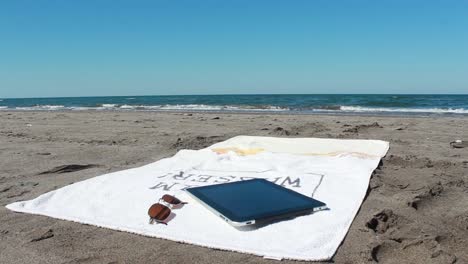 Tableta-Y-Vasos-Sobre-Una-Toalla-Tumbado-En-Una-Playa-Soleada