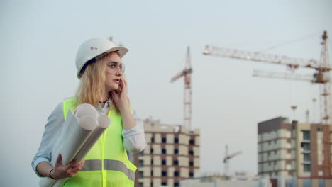 Sprechende-Frau-Mit-Helm-Am-Telefon-Vor-Dem-Hintergrund-Einer-Baustelle-Mit-Kränen,-Die-Zeichnungen-In-Der-Hand-Halten.-Ingenieurin-Auf-Der-Baustelle