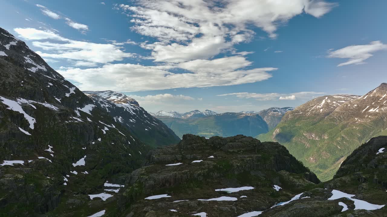 Drone stiger opp bak et granittfjell, og avslører turkise Oldevatnet fjordvann omgitt av snødekte topper under en klar blå sommerhimmel i Norge.