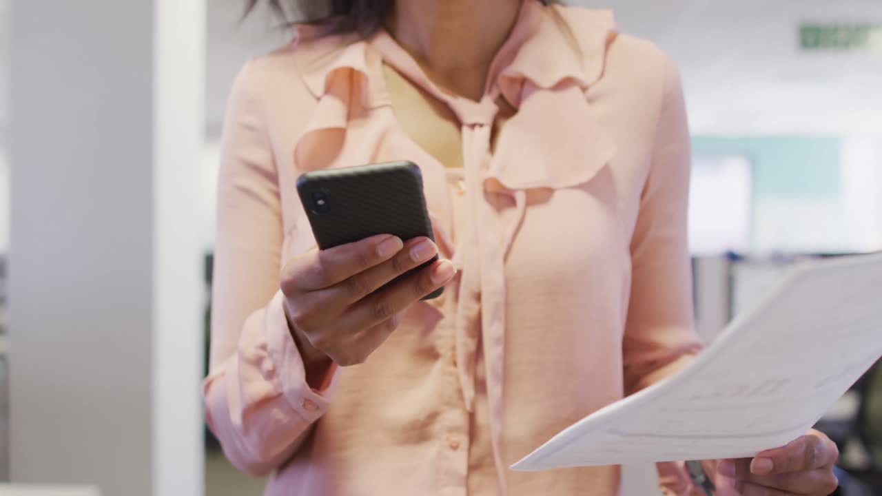 video de la sección media de una mujer de negocios biracial usando un teléfono inteligente y caminando en la oficina