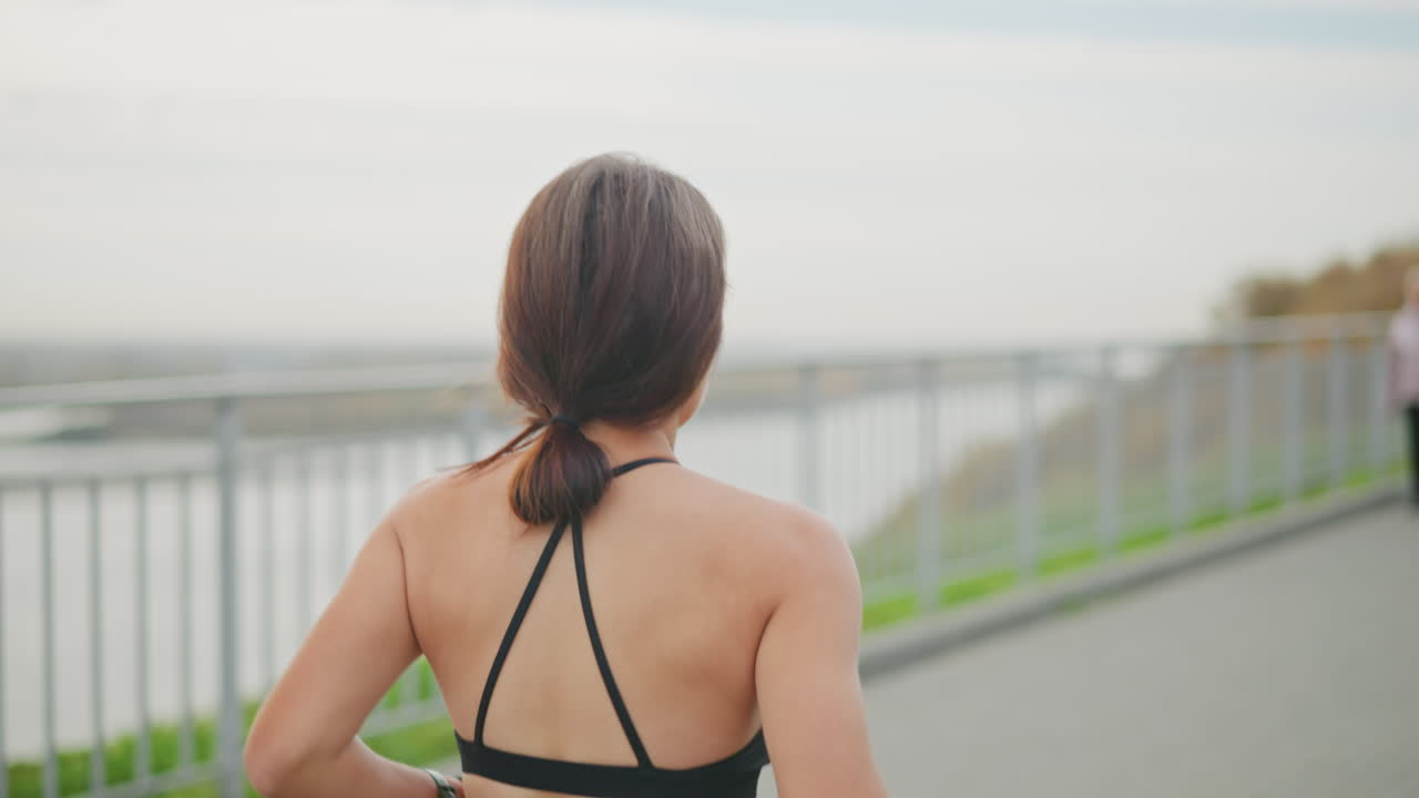 Fair girl in black neck top jogging beside iron fence with blurred view of a woman walking past her in park, capturing outdoor fitness, health, and motion in bright surroundings