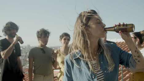 portrait of a young caucasian woman looking at the camera and drinking beer while having a rooftop party with friends