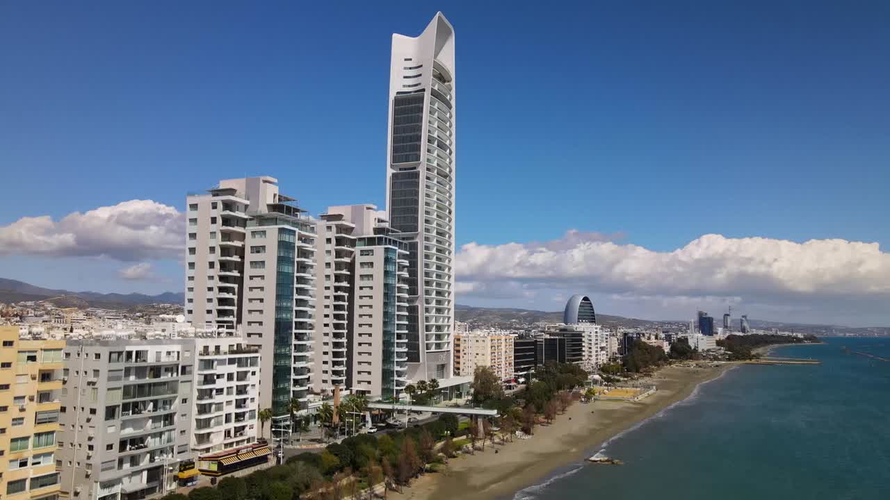 Drone footage of modern skyscraper lining Sarandë's picturesque waterfront with sandy beach, mountain backdrop. Sunny, vibrant views showcase urban landscape and coastal charm