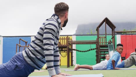 Diverse-male-teacher-and-happy-schoolchildren-exercising-on-mats-at-school-playground