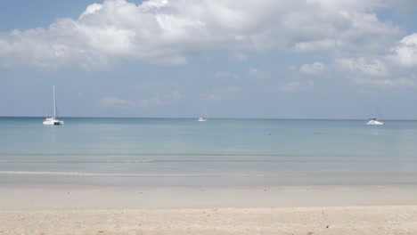 Der-Strand-Mit-Ein-Paar-Yachten-Im-Meer,-Weißer-Sand-Und-Wellen-Vom-Ruhigen-Meer-Bei-Sonnenschein-Tagsüber