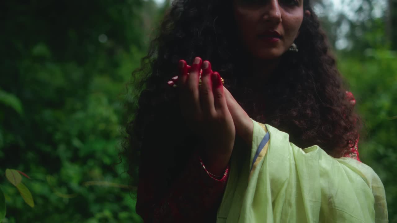 Close-up of woman with curly hair holding her hands together, wearing a green sari, surrounded by lush vegetation