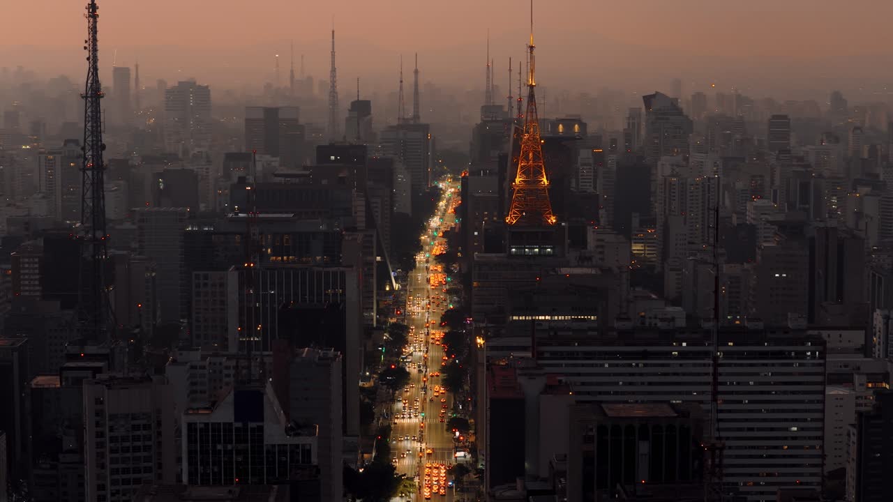 Panoramic aerial view panning left over Av. Paulista during sunset, with golden-lit communications tower and traffic lights glowing across the skyline.