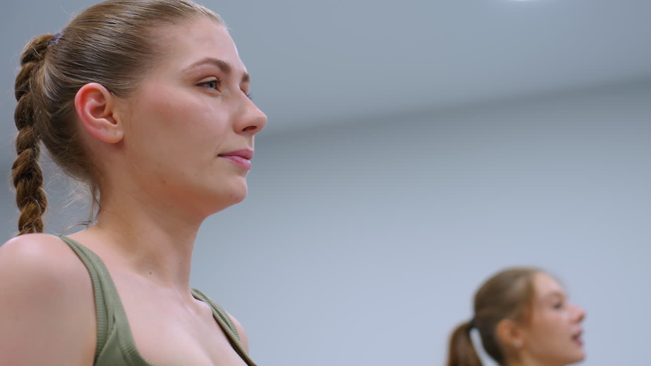 Close-up of women working out, rising from squats, taking deep breaths as bright lighting illuminates the scene, focusing on their concentration and controlled movements in a fitness session