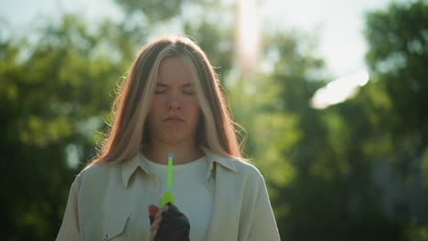 chica de pelo rubio de pie con una expresión pensativa, examinando cuidadosamente la boquilla de la bomba de aire verde contra el fondo de árboles exuberantes y iluminados por el sol