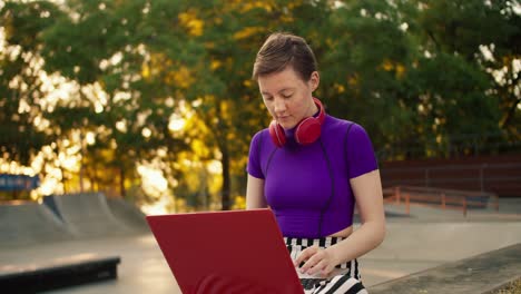 Portrait-of-a-young-short-haired-girl-in-a-purple-swamp-who-sits-on-the-street-in-red-headphones-and-works-on-a-Red-laptop-in-summer