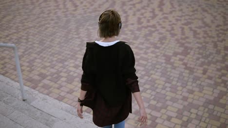 Happy-stylish-woman-with-ponytail-going-down-the-stairs-playfully-in-the-park.-Whirling-and-dancing-with-music-in-her-headphones