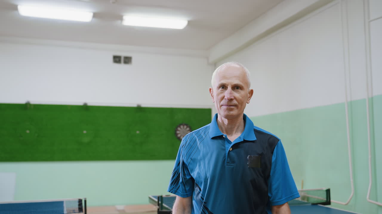 Old man wearing blue sports shirt folding hands and posing confidently in front of camera indoors near table tennis tables, expressing calm attitude