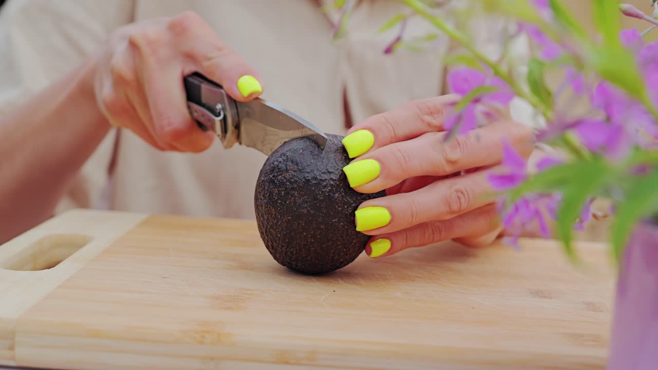 Woman’s bright nails slice avocado skin slowly at rustic campsite table