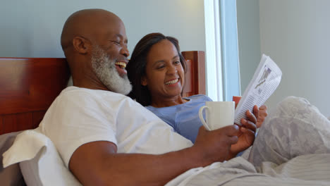 Side-view-of-mixed-race-mature-couple-reading-newspaper-and-drinking-coffee-in-bedroom-at-home-4k