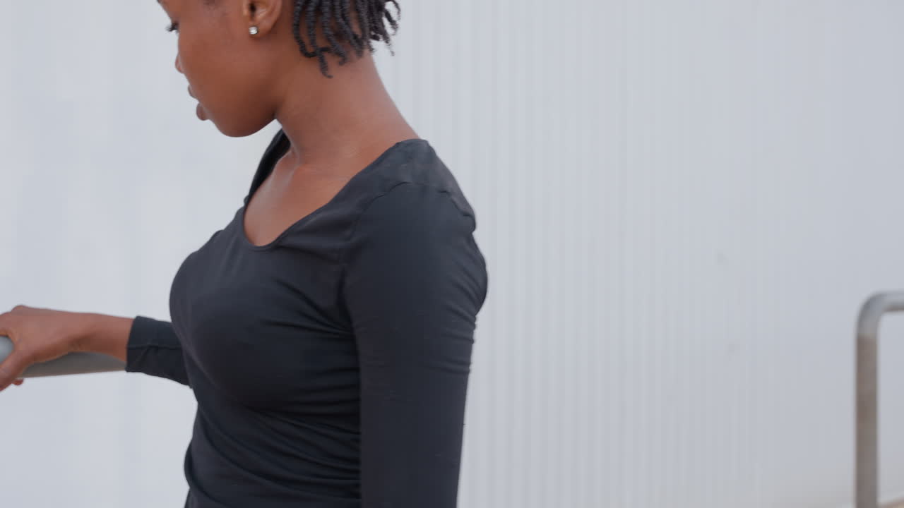woman leaning on metal railing, contemporary dancer warming up, braided hair, black top, focused downward gaze, controlled shoulder stretch, industrial backdrop, quiet mood, soft neutral tones