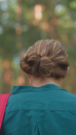 vista trasera de una mujer caminando por un pacífico bosque iluminado por el sol con una mochila roja colgada sobre su hombro, el cabello cuidadosamente atado hacia atrás, la luz del sol se filtra a través de árboles altos