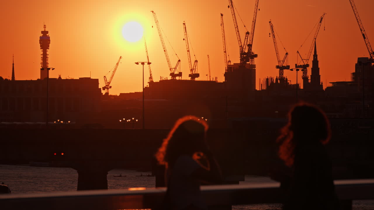 Two women standing by the railing watching a vibrant sunset over cranes and the London skyline, London, England