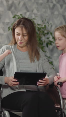 mother smiles showing videos to curious children on tablet. toddler and preschooler stand near mommy with disability sitting in wheelchair in room