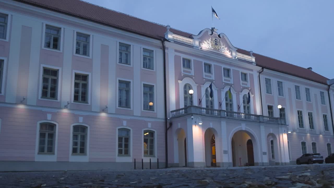 Footage shows Estonian government parliament building during an autumn evening or night while the building is lit with lights. An Estonian flag is waving in slow motion on the top and clouds visible.
