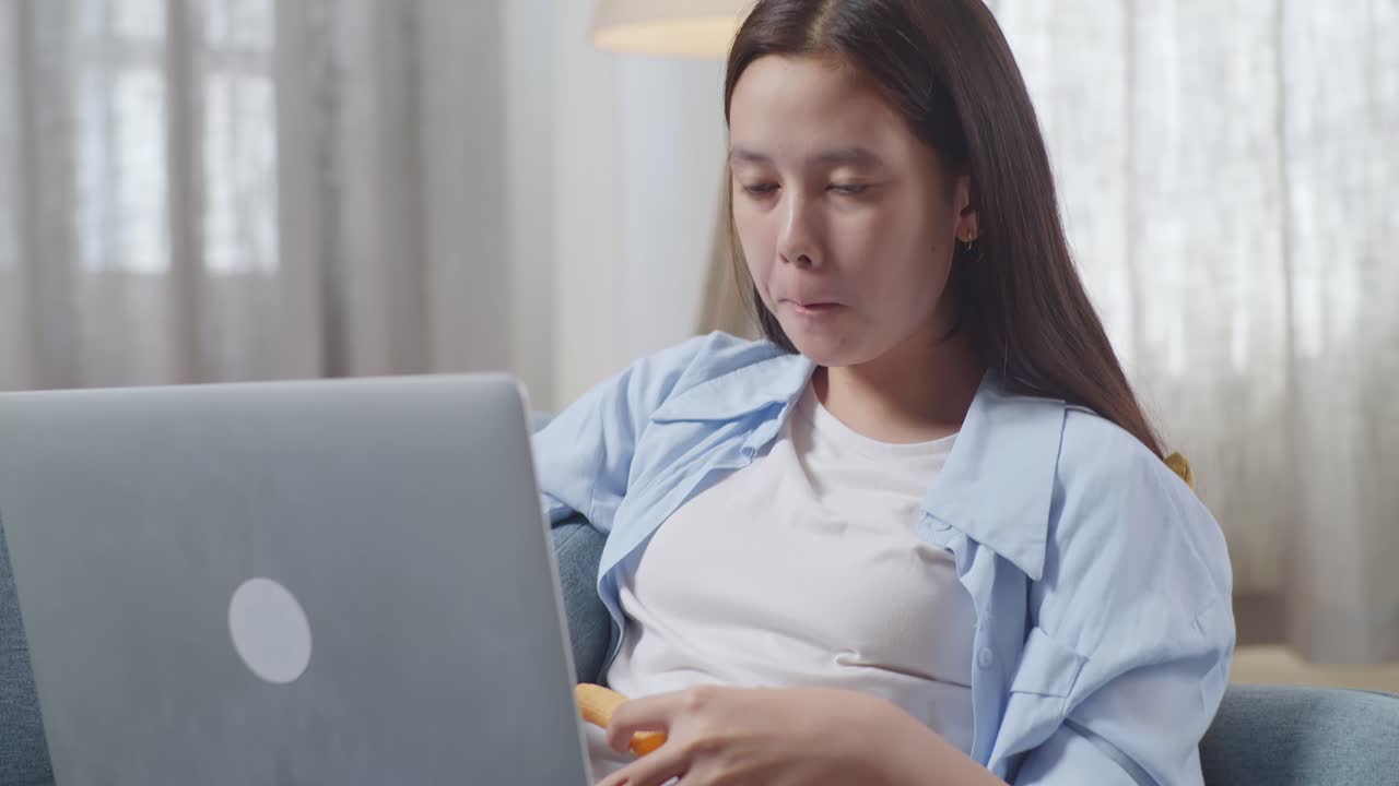 아시안 십대 소녀가 집에서 거실에서 소파에 누워서 간식을 먹고 노트북으로 타이핑하는 장면