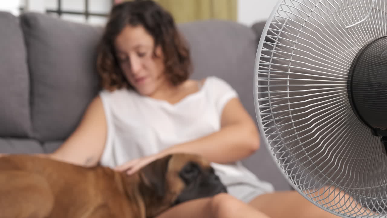 young woman petting her boxer dog on the sofa, with the fan on