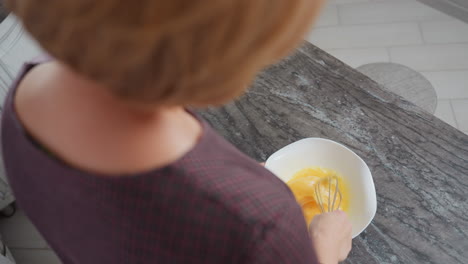 rear right side view of girl with golden hair wearing plaid gown stirring egg in bowl, she lifts whisk briefly before continuing mixing, with a sleek marble countertop and light-colored tiled floor