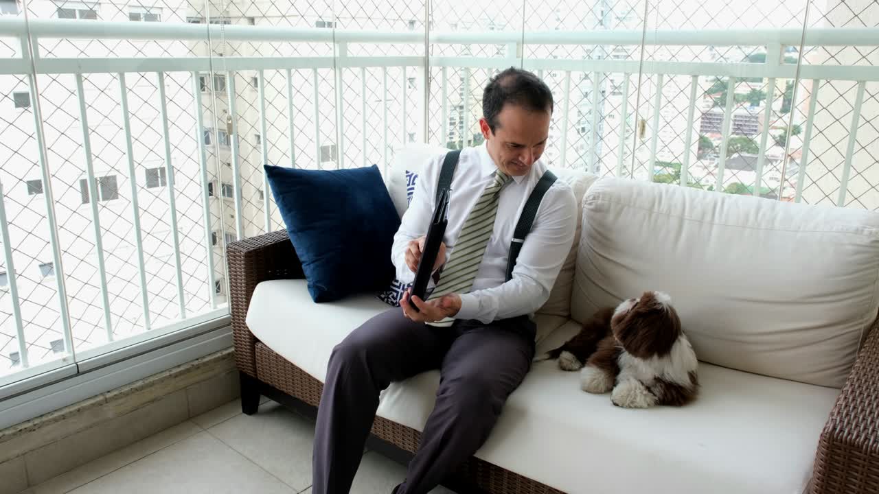 타이와 서펜더를 입은 남자가 소파에 앉아서 그의 애완동물 (시츠) 에 있는 태블릿으로 비디오 통화를 하고 있습니다.