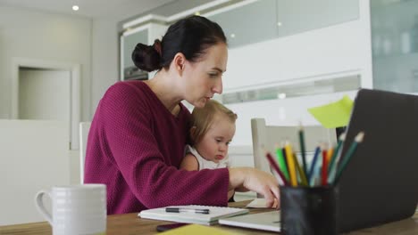 caucasian mother holding her baby using laptop while working from home