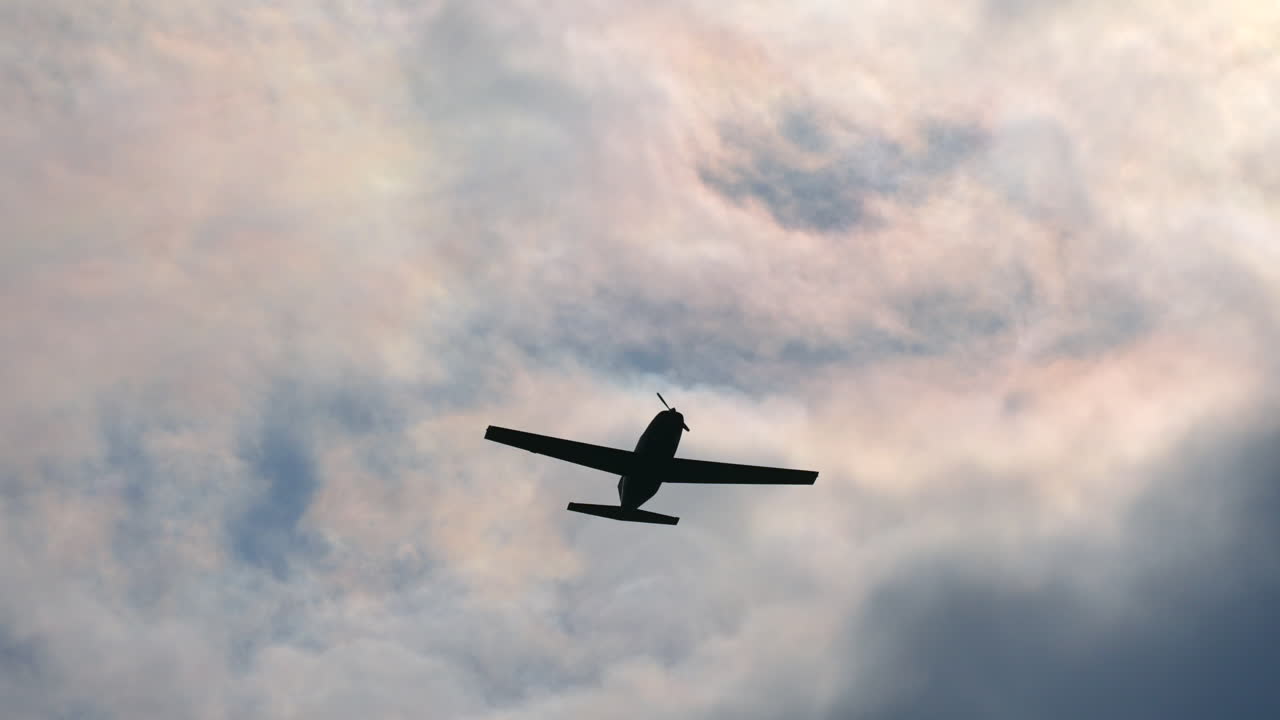 Tracking shot showing silhouette of airplane flying against sunlight and dark clouds at sky.