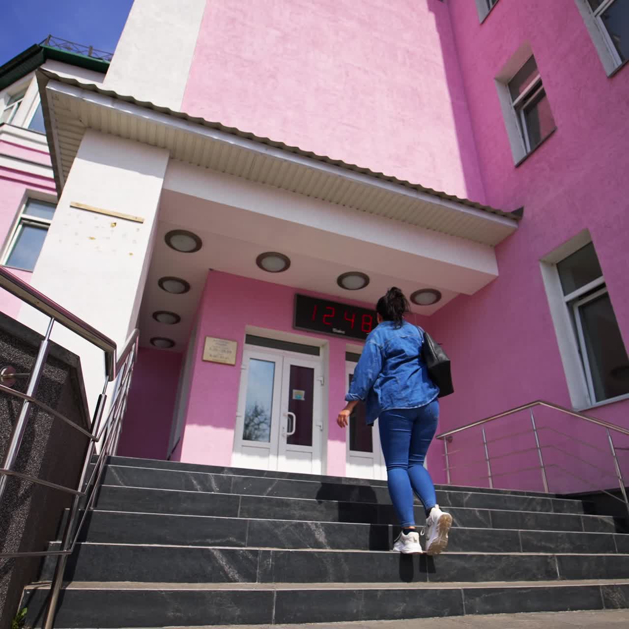 Coming up by the stairs with silver handrails to the doors of the big building. Following the girl in jeans wear going up by the staircase. Low angle view