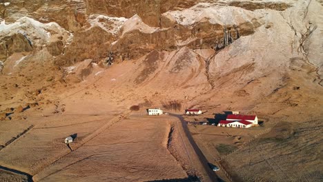 Luftaufnahme-Einer-Kargen-Landschaft-An-Der-Südküste-Islands,-Mit-Ein-Paar-Häusern-Mit-Roten-Dächern-Auf-Einer-Großen-Fläche-Land-Vor-Der-Kulisse-Hoher-Berge,-Die-In-Goldenes-Licht-Getaucht-Sind