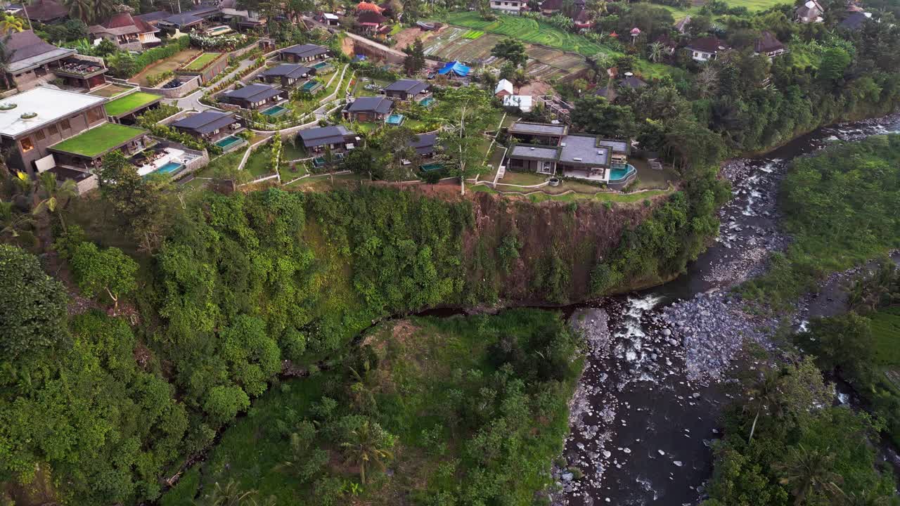 Drone footage of modern villas built along a green cliff above the river in Sidemen, Bali. The aerial view highlights the mix of tropical vegetation, contemporary design, and peaceful rural landscape