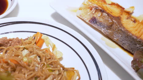 panning from the right to the left on a full serving of meal consisting of salmon steak, stir-fried noodles, and some salad greens