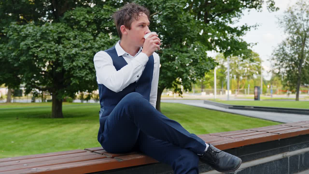Young caucasian man in a suit vest having a pensive coffee break, sitting alone on a park bench, reflecting and enjoying a moment of solitude outdoors, looking away, slow motion push in shot