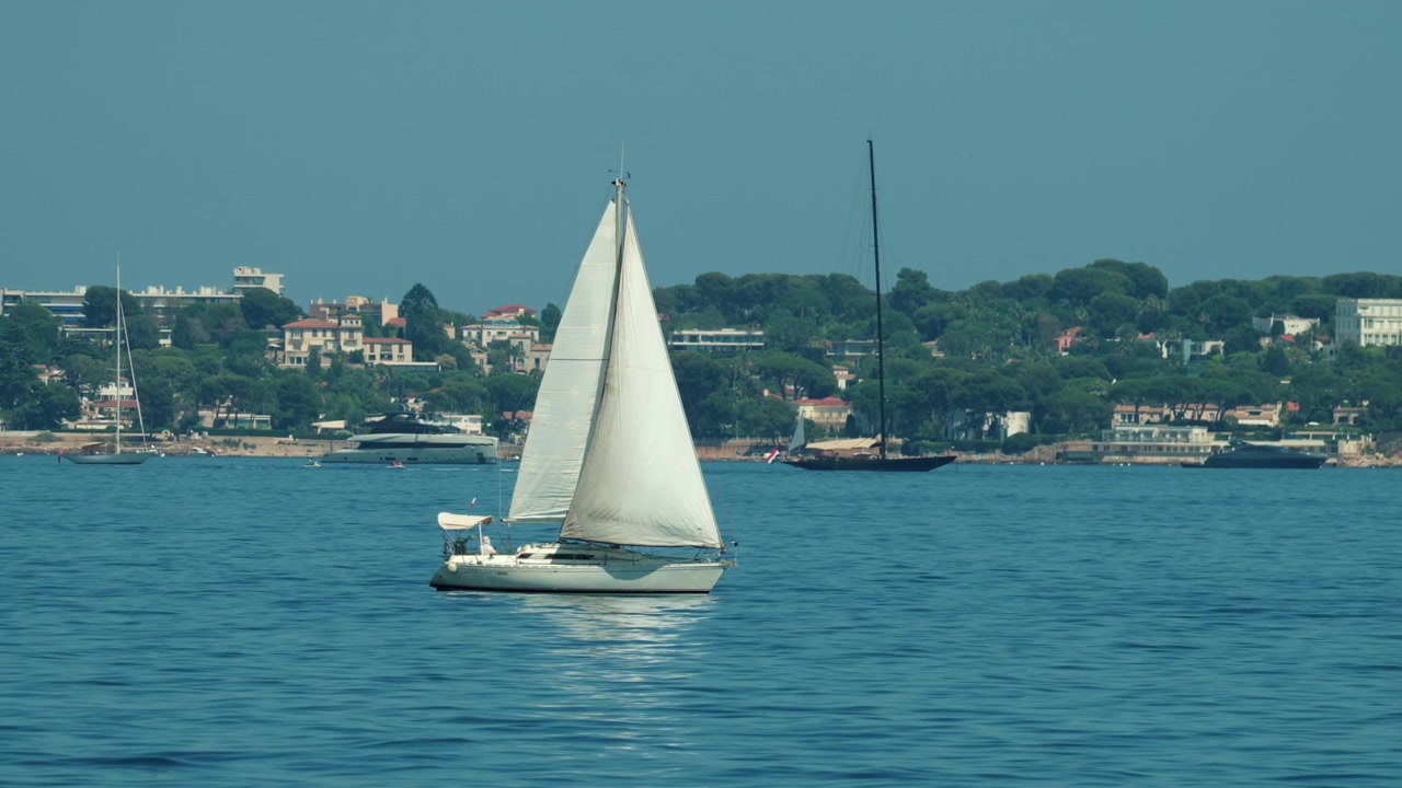 Berrak bir gökyüzü altında, arka planda yemyeşil bir sahil kasabasıyla mavi sakin sularda seyreden beyaz yelkenli, Golfe Juan, Fransa.