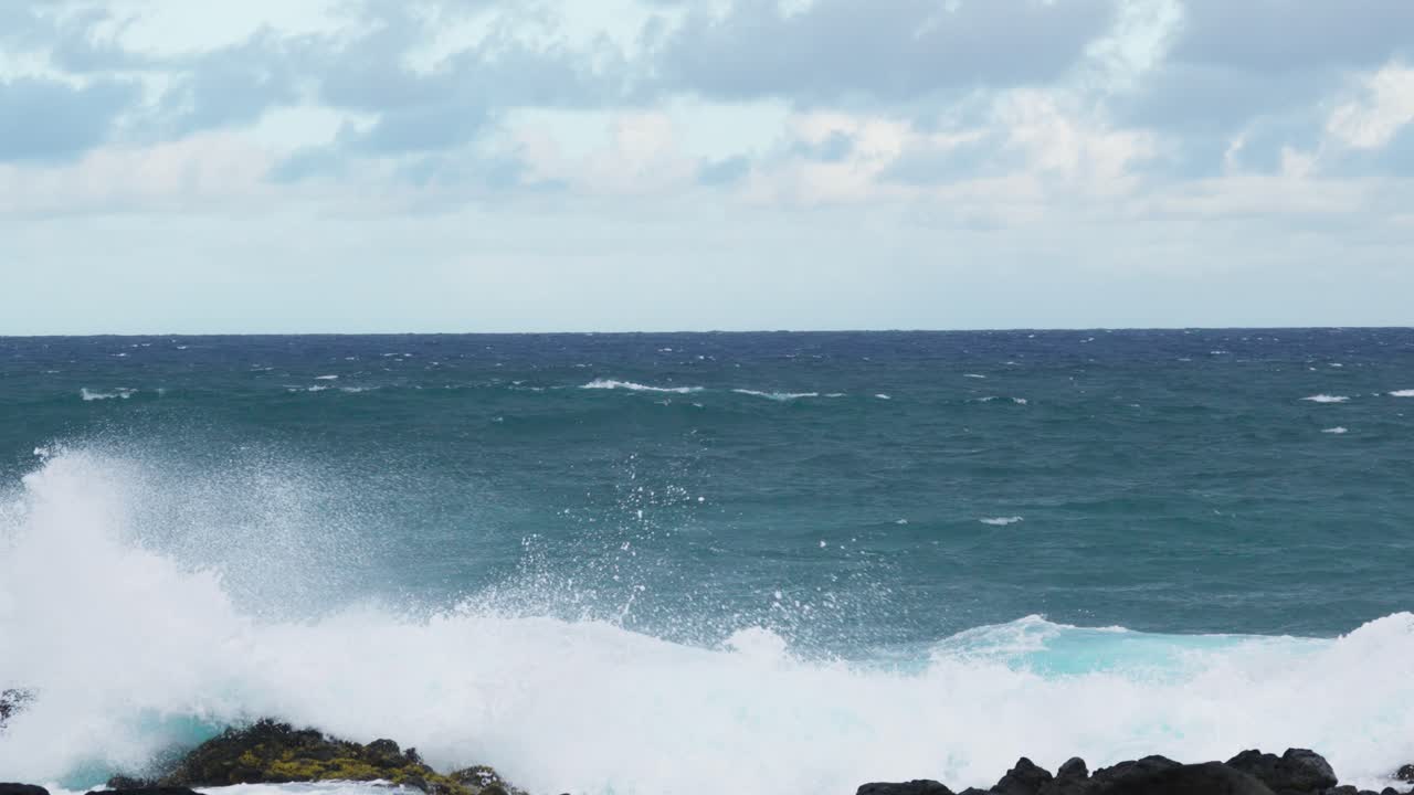 Wide view of turquoise ocean waves colliding with dark jagged rocks, foamy white surf splashing upward against the rugged coastline beneath a cloudy horizon