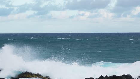 wide view of turquoise ocean waves colliding with dark jagged rocks, foamy white surf splashing upward against the rugged coastline beneath a cloudy horizon
